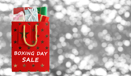 Image of Red shopping bag with text Boxing Day Sale full of gifts on blurred silver background, closeup. Space for text