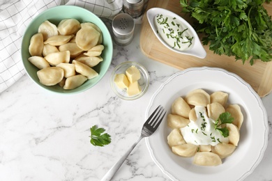 Photo of Delicious cooked dumplings with sour cream on white marble table, flat lay