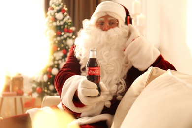 Photo of MYKOLAIV, UKRAINE - JANUARY 18, 2021: Santa Claus listening to music with headphones at home, focus on Coca-Cola bottle in his hand