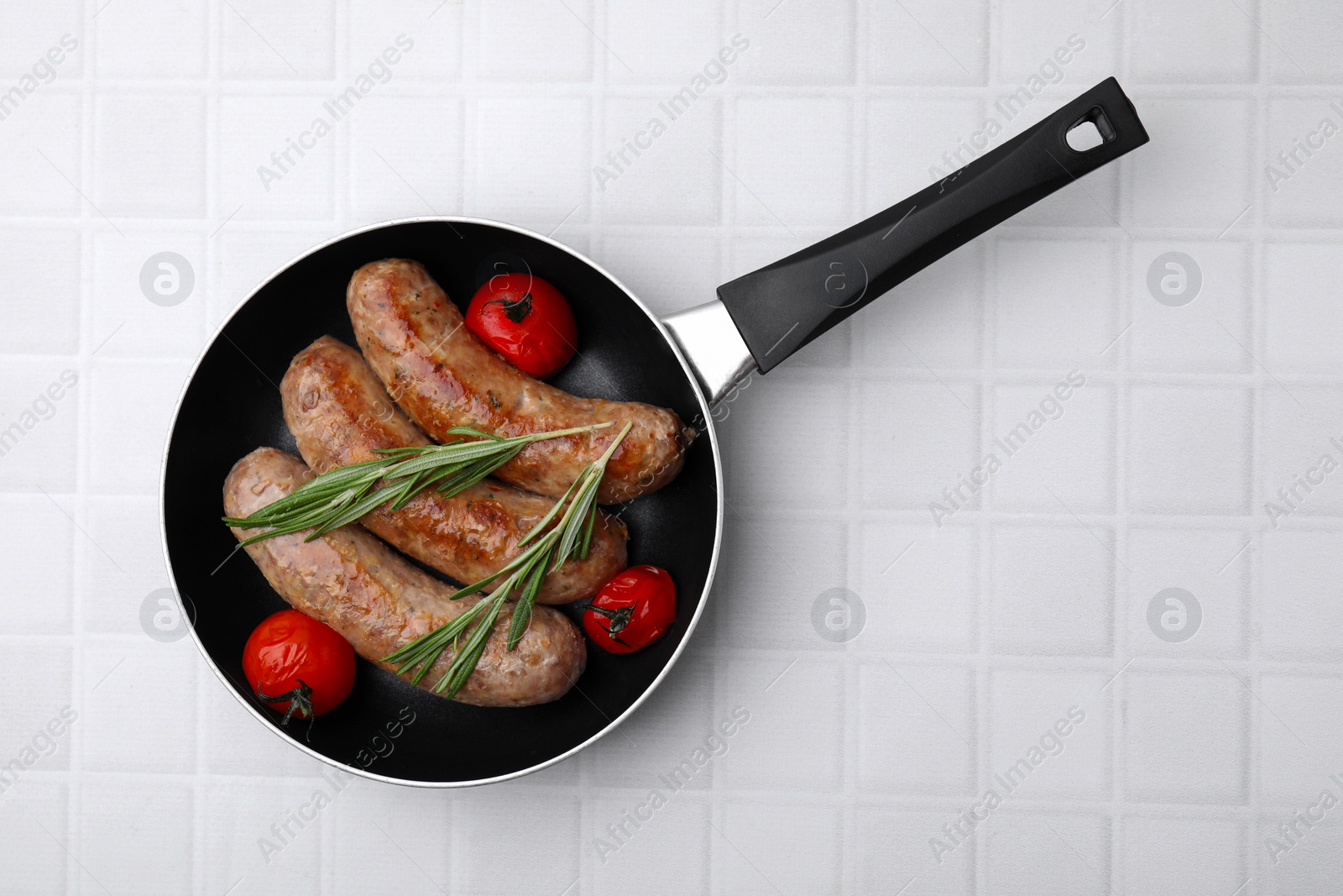 Photo of Frying pan with tasty homemade sausages, rosemary and tomatoes on white tiled table, top view