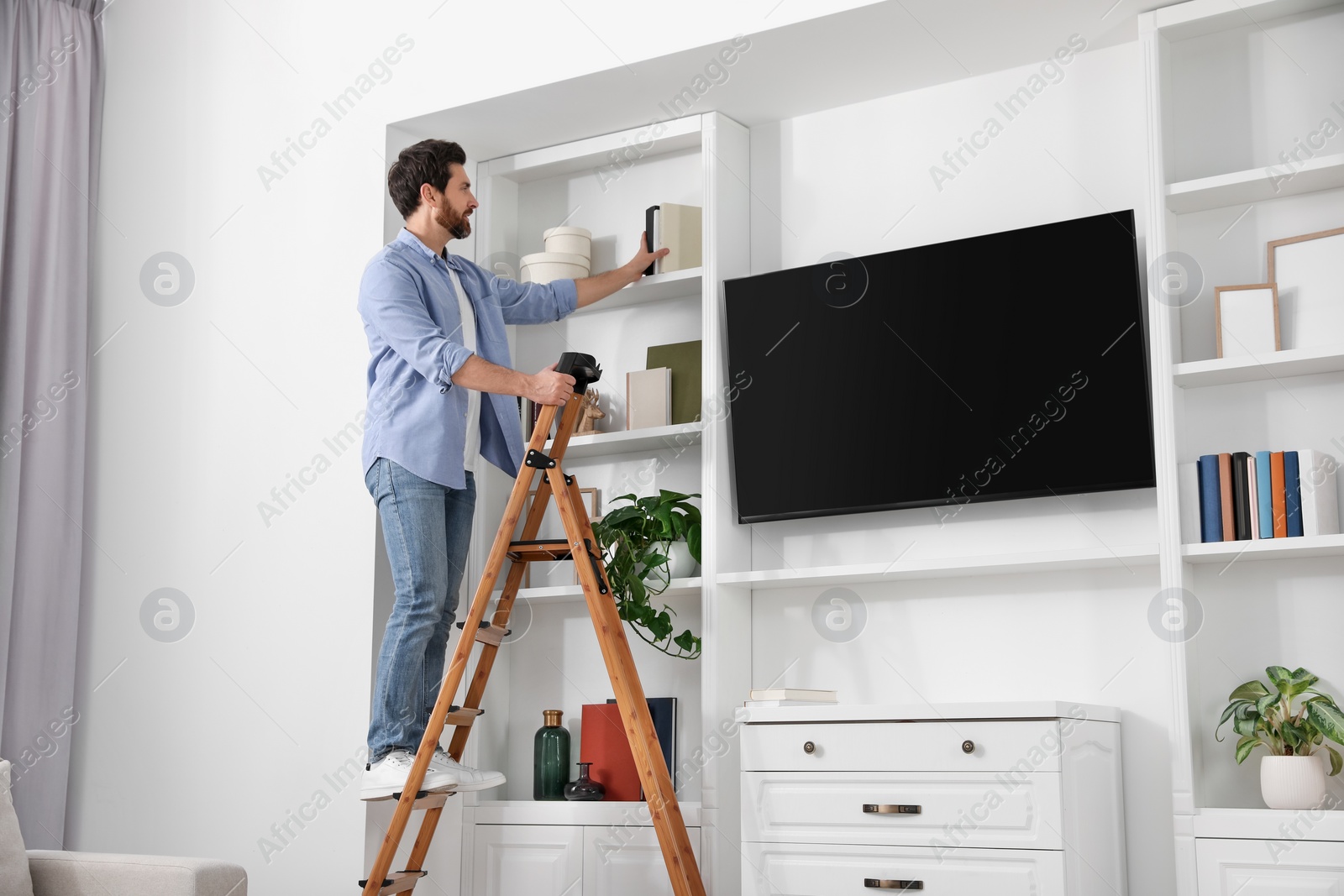 Photo of Man on wooden folding ladder taking book from shelf at home