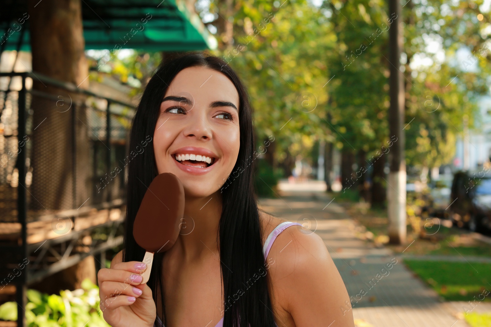 Photo of Beautiful young woman eating ice cream glazed in chocolate on city street