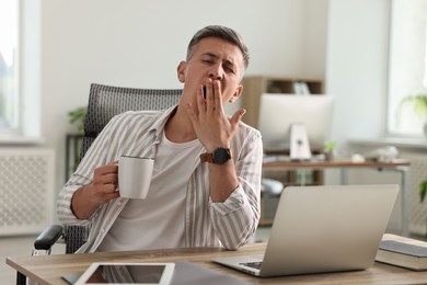 Photo of Man with cup of drink yawning at wooden table in office