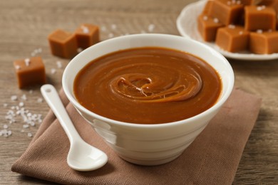 Photo of Yummy salted caramel in bowl and candies on wooden table, closeup