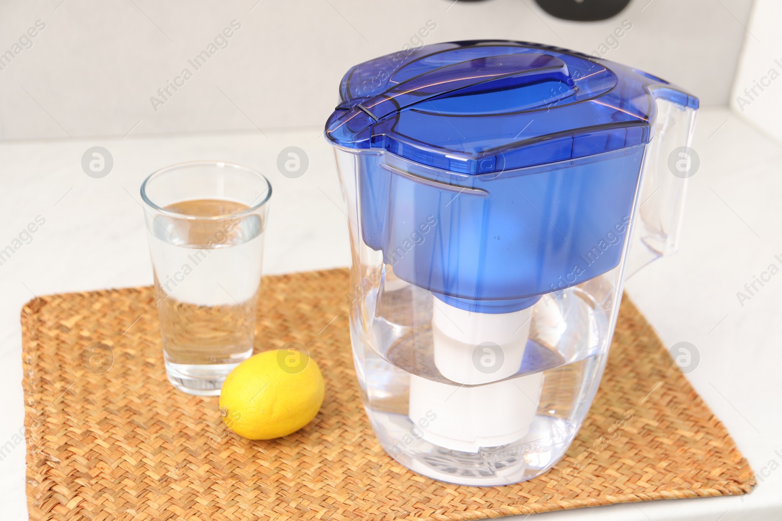 Photo of Water filter jug, glass and lemon on white countertop in kitchen