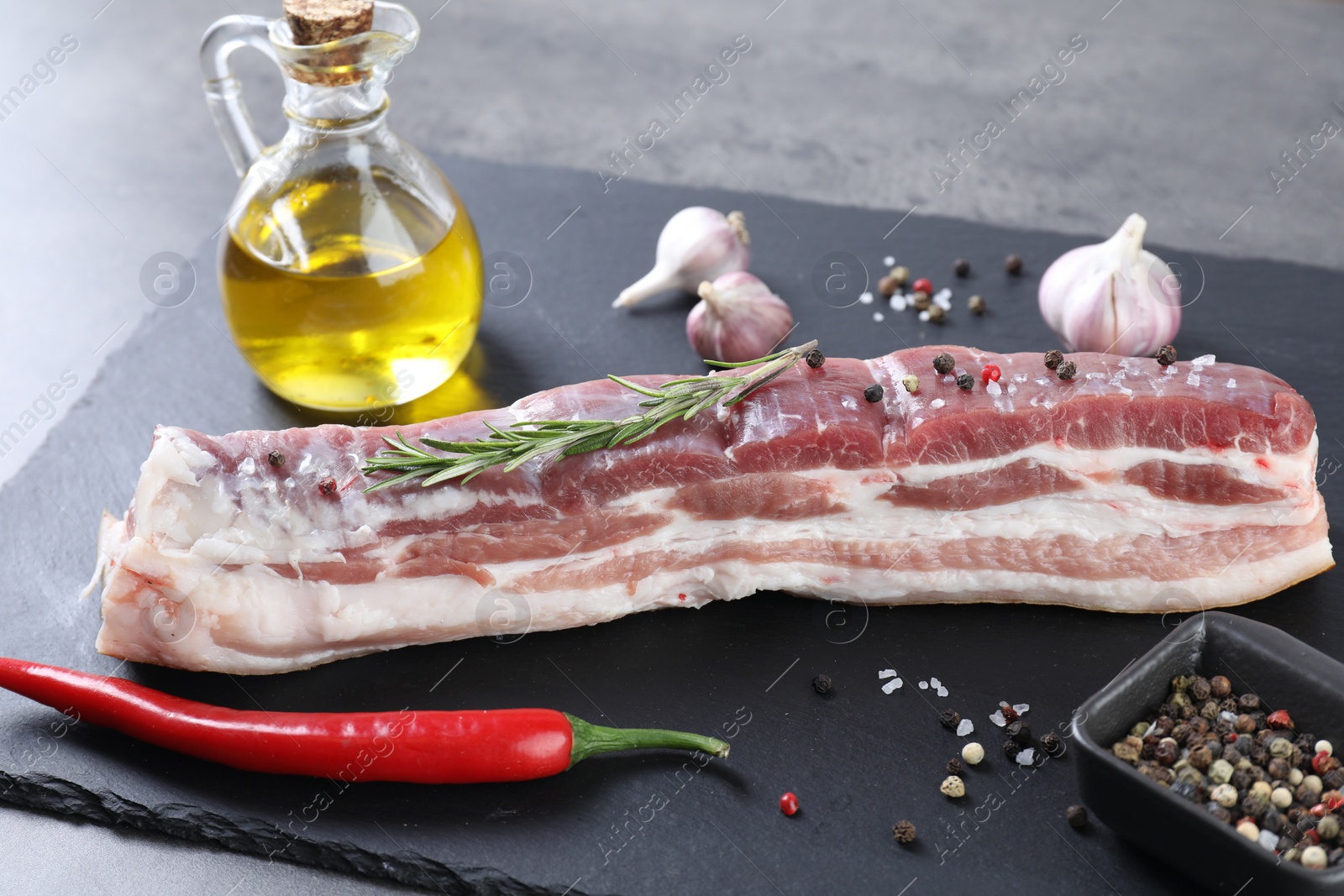 Photo of Piece of raw pork belly, rosemary, chili pepper and spices on grey table