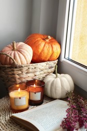 Beautiful heather flowers, burning candles, open book and wicker basket with pumpkins near window indoors