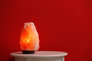 Photo of Himalayan salt lamp on table against dark red background. Space for text