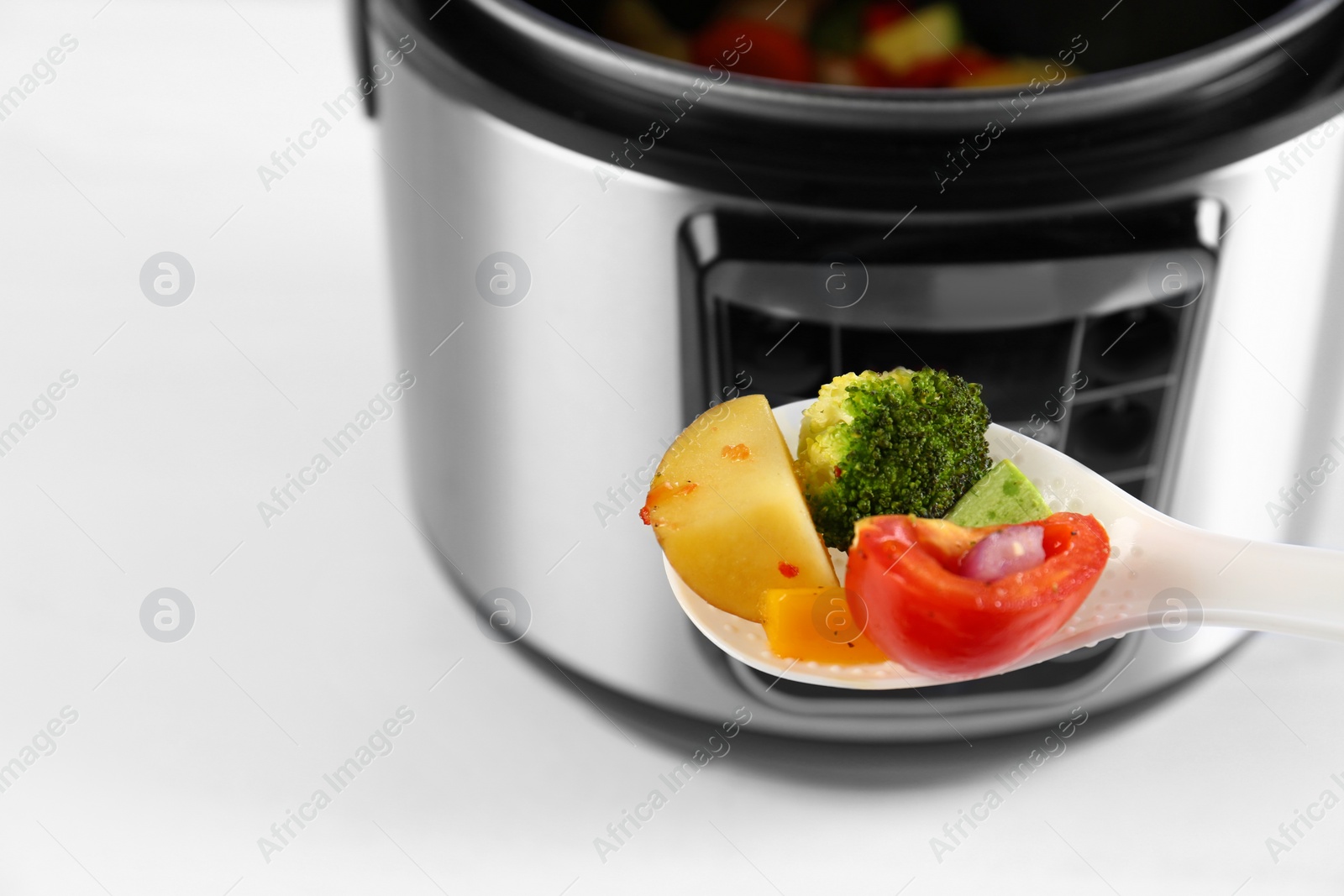 Photo of Spoon with vegetables and blurred view of modern multi cooker on background, closeup. Space for text