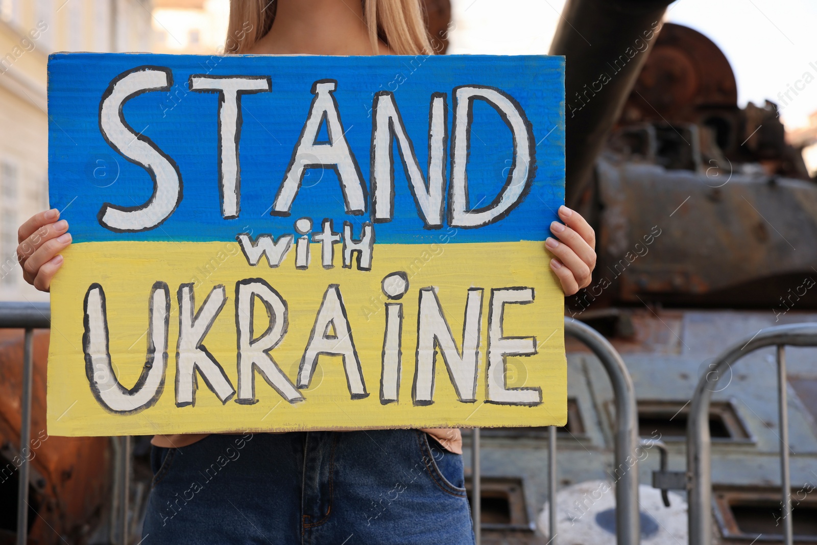Photo of Woman holding poster in colors of national flag with words Stand with Ukraine near broken tank on city street, closeup