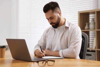 Photo of Young man writing down notes during webinar at table in room