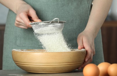 Photo of Woman sieving flour into bowl at table in kitchen, closeup