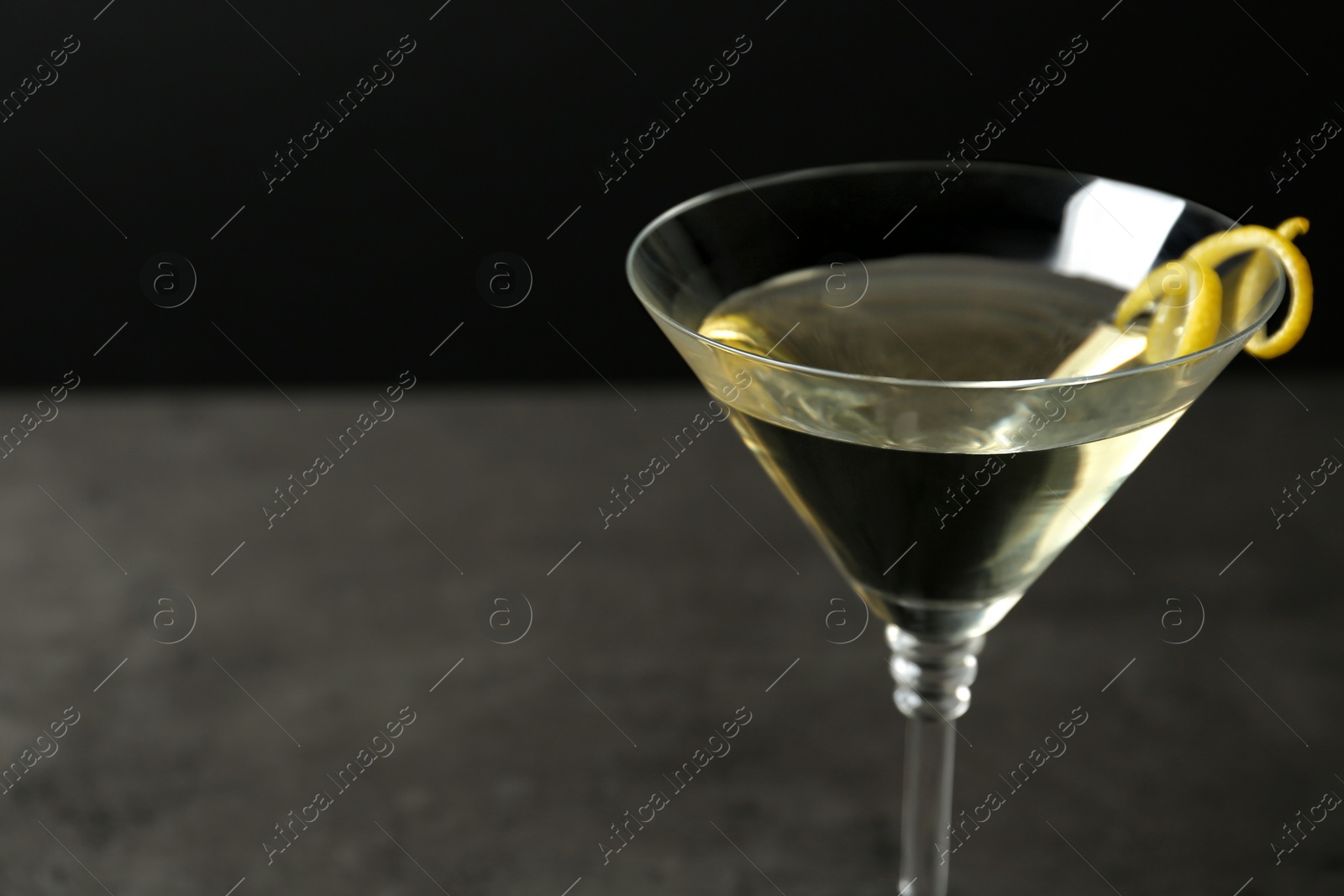 Photo of Glass of lemon drop martini cocktail with zest on stone table against black background, closeup. Space for text
