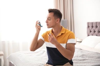 Young man with asthma inhaler on bed in light room