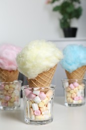 Photo of Sweet cotton candies in waffle cones and marshmallows on white table indoors, closeup