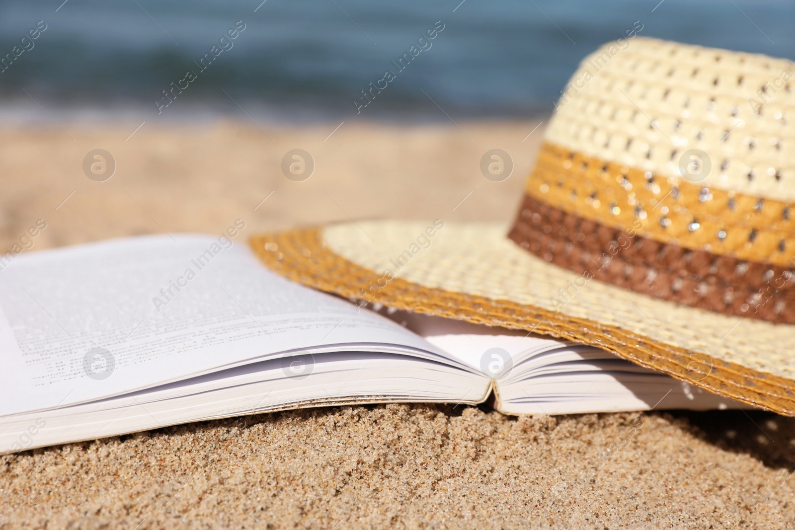 Photo of Open book and hat on sandy beach near sea, closeup