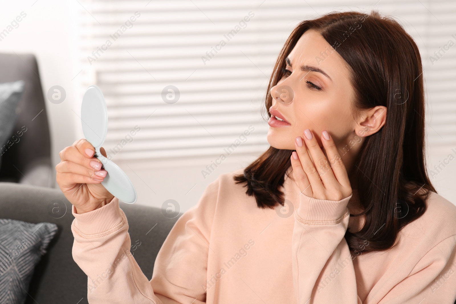 Photo of Young woman looking in mirror and touching her face indoors. Hormonal disorders