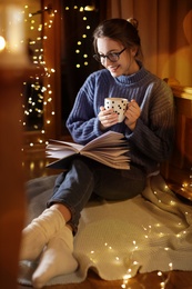 Woman with cup of hot beverage reading book at home in winter evening