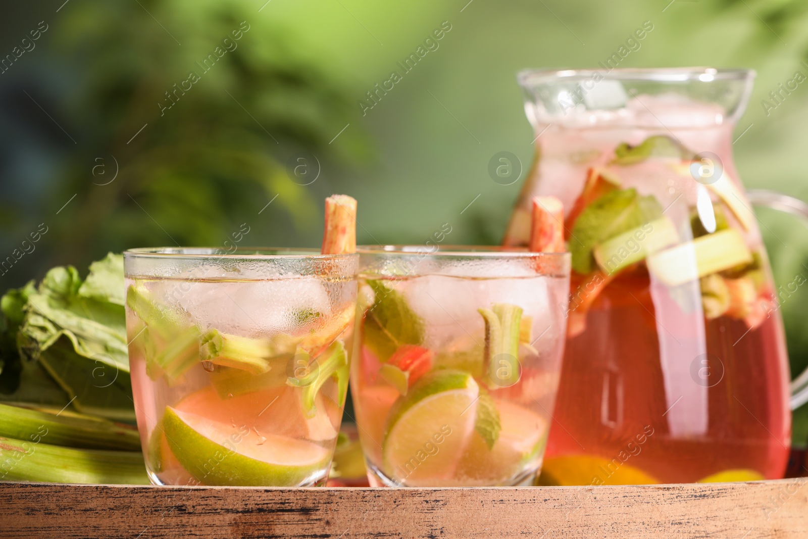 Photo of Glasses and jug of tasty rhubarb cocktail with citrus fruits on wooden board outdoors, closeup