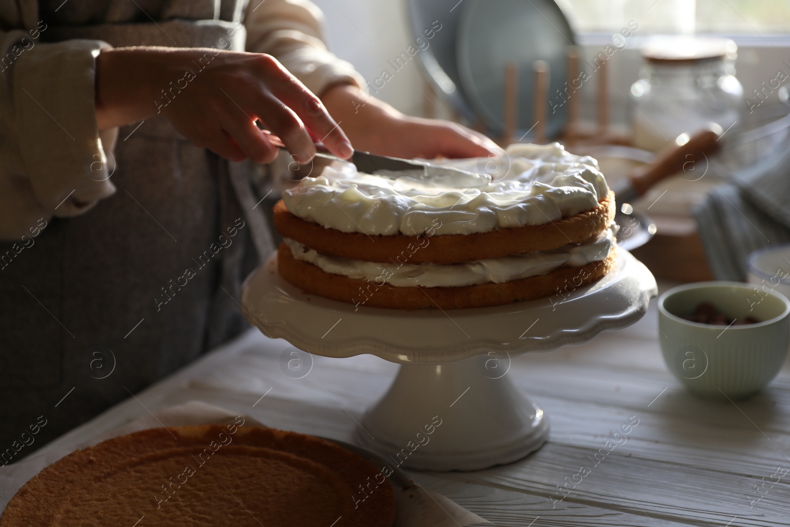 Photo of Woman smearing sponge cake with cream at white wooden table in kitchen, closeup