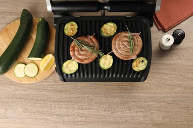 Photo of Electric grill with homemade sausages, zucchini and rosemary on wooden table, flat lay. Space for text