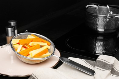 Photo of Sieve with cut raw parsnips and carrots on black table in kitchen