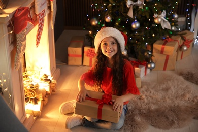 Photo of Cute little child in Santa hat with Christmas gift sitting in front of fireplace near fir tree at home