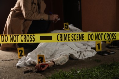 Photo of Investigator examining crime scene with dead body outdoors, focus on yellow tape