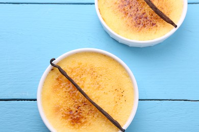Photo of Delicious creme brulee in bowls and vanilla pods on light blue wooden table, top view