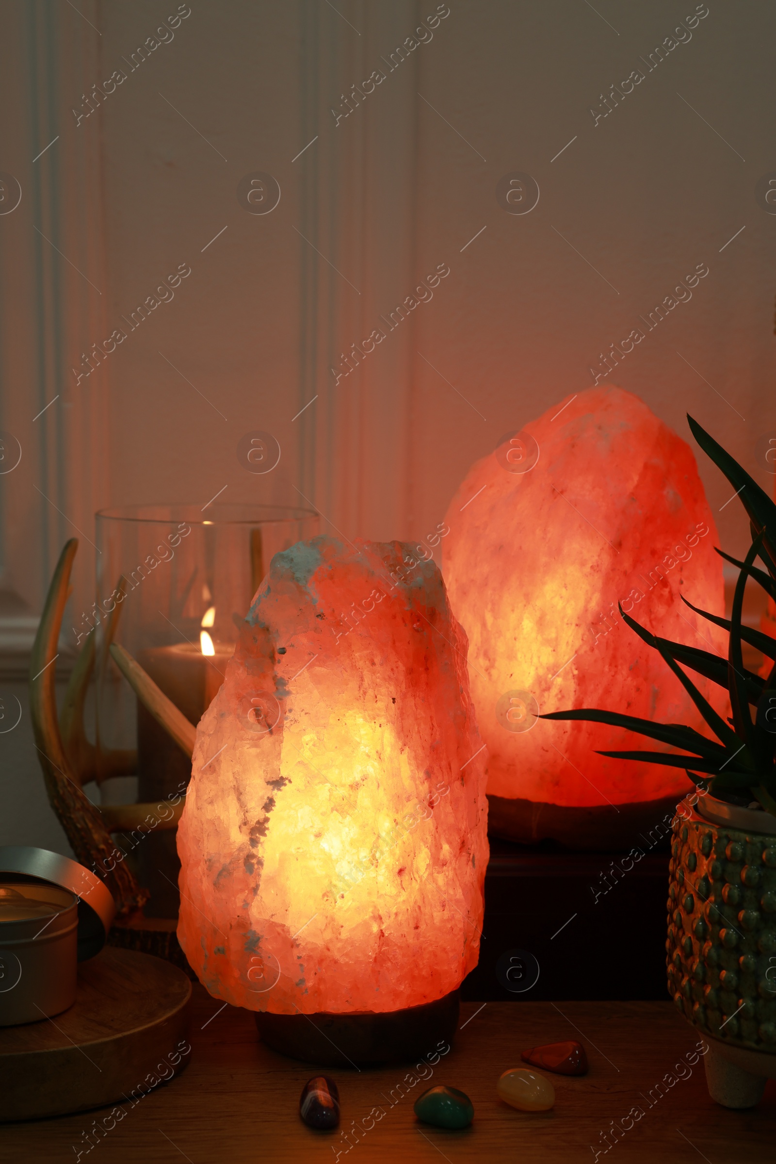 Photo of Himalayan salt lamps, candle, houseplant and gemstones on wooden table near white wall indoors