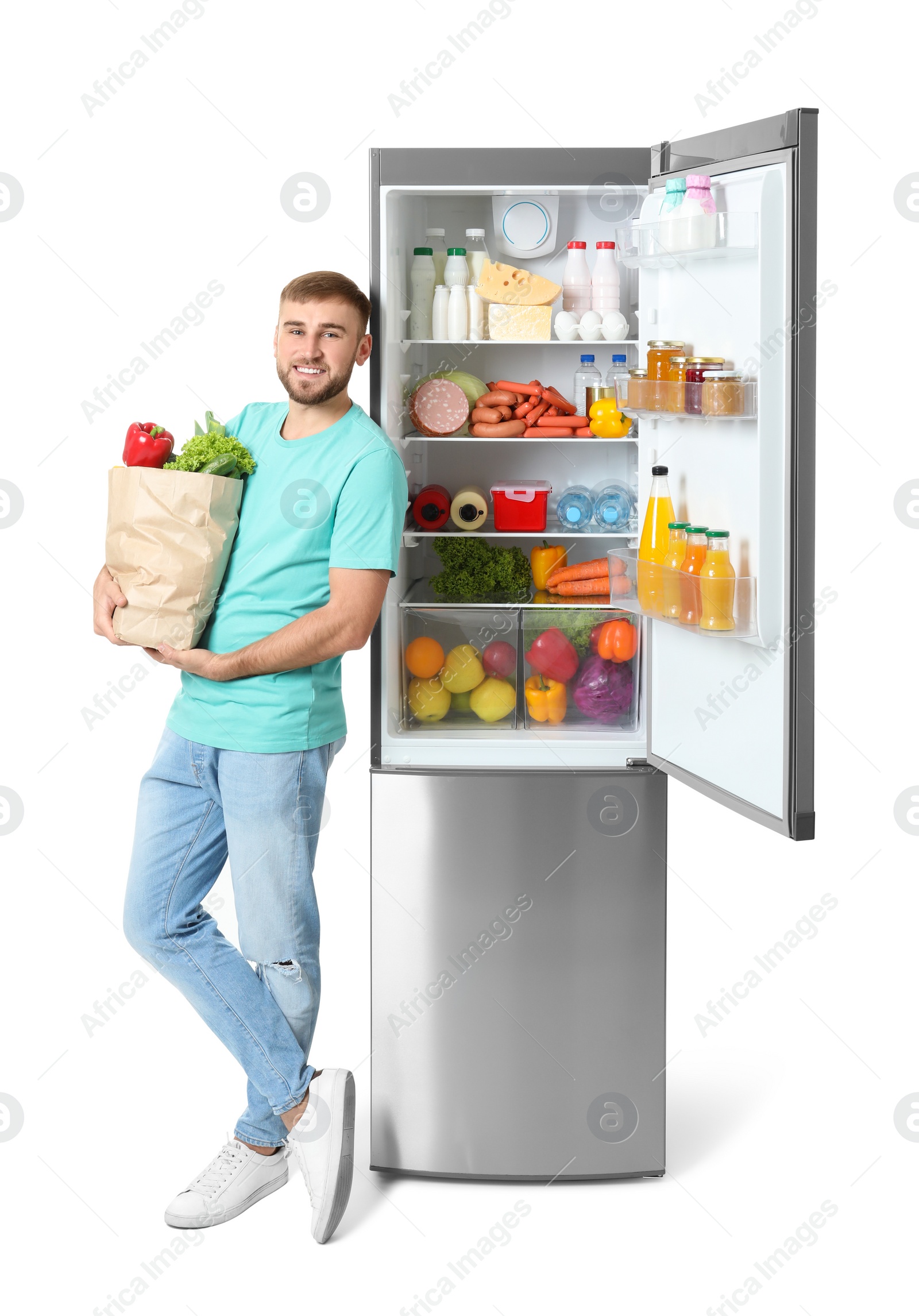 Photo of Young man with bag of groceries near open refrigerator on white background