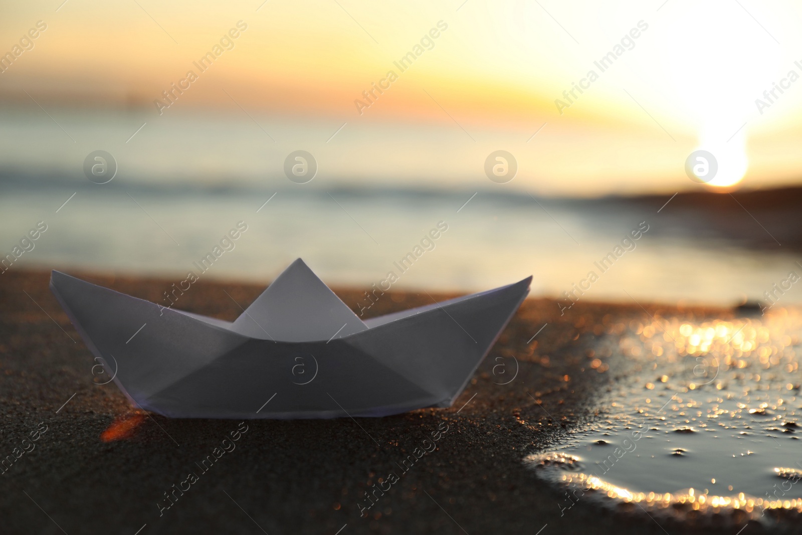 Photo of White paper boat on sand near sea at sunset, closeup. Space for text