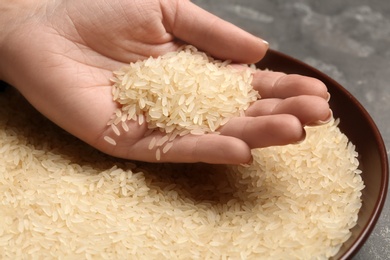 Woman holding grains near plate with parboiled rice on table, closeup