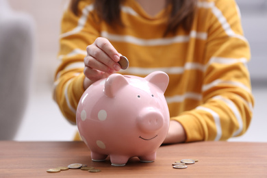 Photo of Woman putting coin into piggy bank at wooden table, closeup