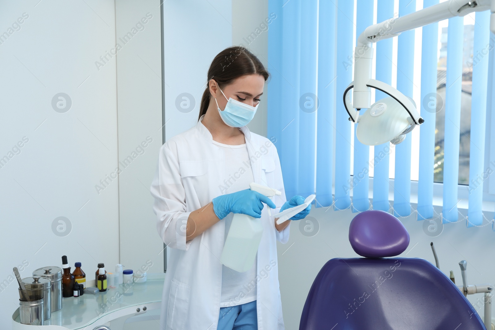 Photo of Professional dentist in white coat and medical mask cleaning workplace with antiseptic indoors