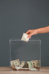 Woman putting money into donation box on table against grey background, closeup