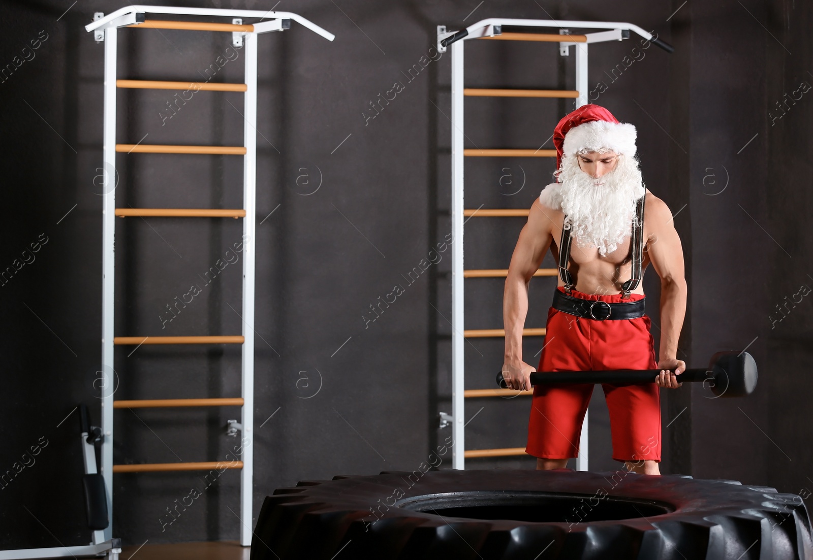 Photo of Young muscular man in Santa costume hitting heavy tire with hammer at gym