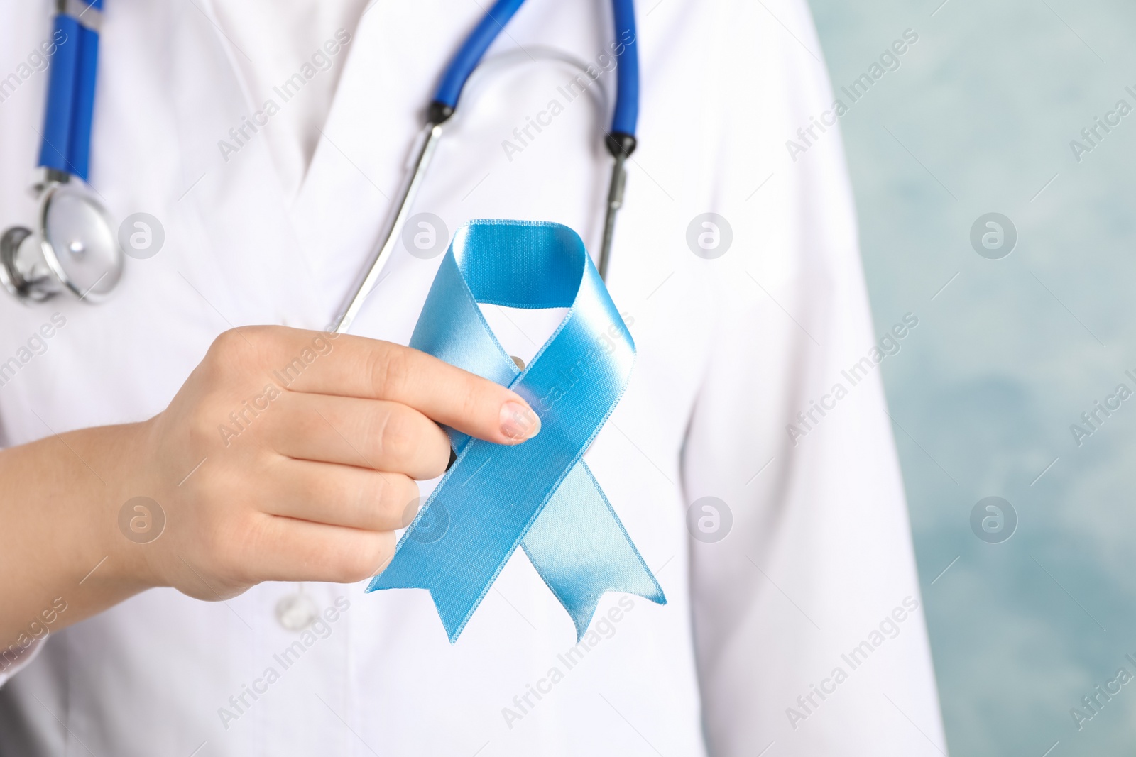 Photo of Doctor holding light blue awareness ribbon on color background, closeup