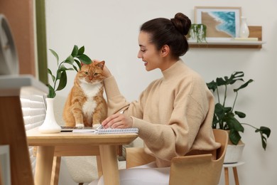 Photo of Woman writing in notebook and petting cute cat at home