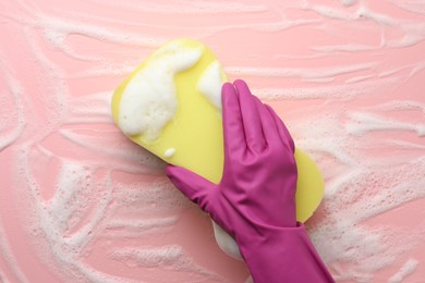 Photo of Cleaner in rubber glove holding sponge with foam on pink background, top view.