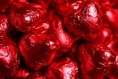 Photo of Tasty chocolate heart shaped candies in red foil wrappers as background, closeup