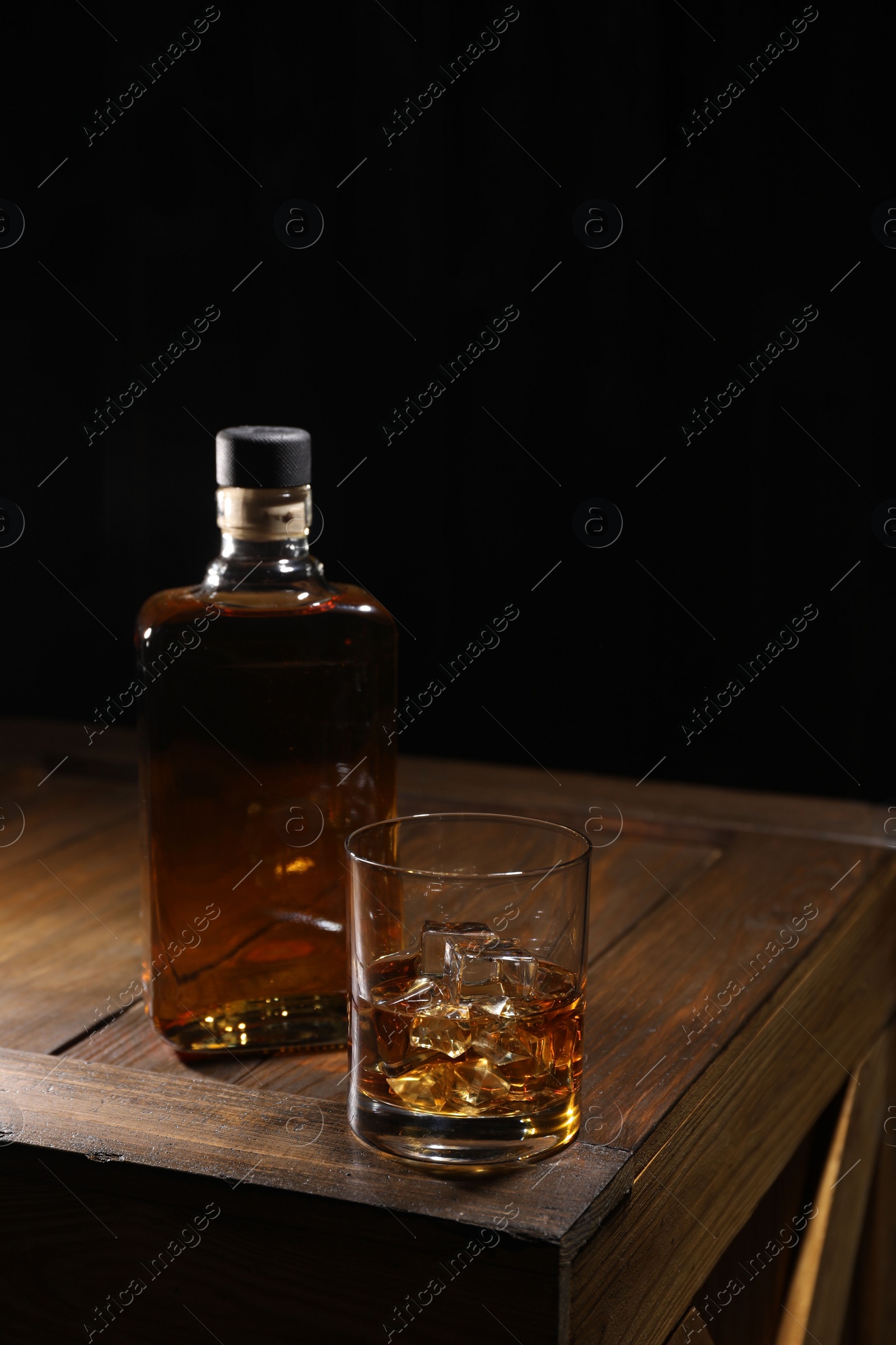 Photo of Whiskey with ice cubes in glass and bottle on wooden crate against black background