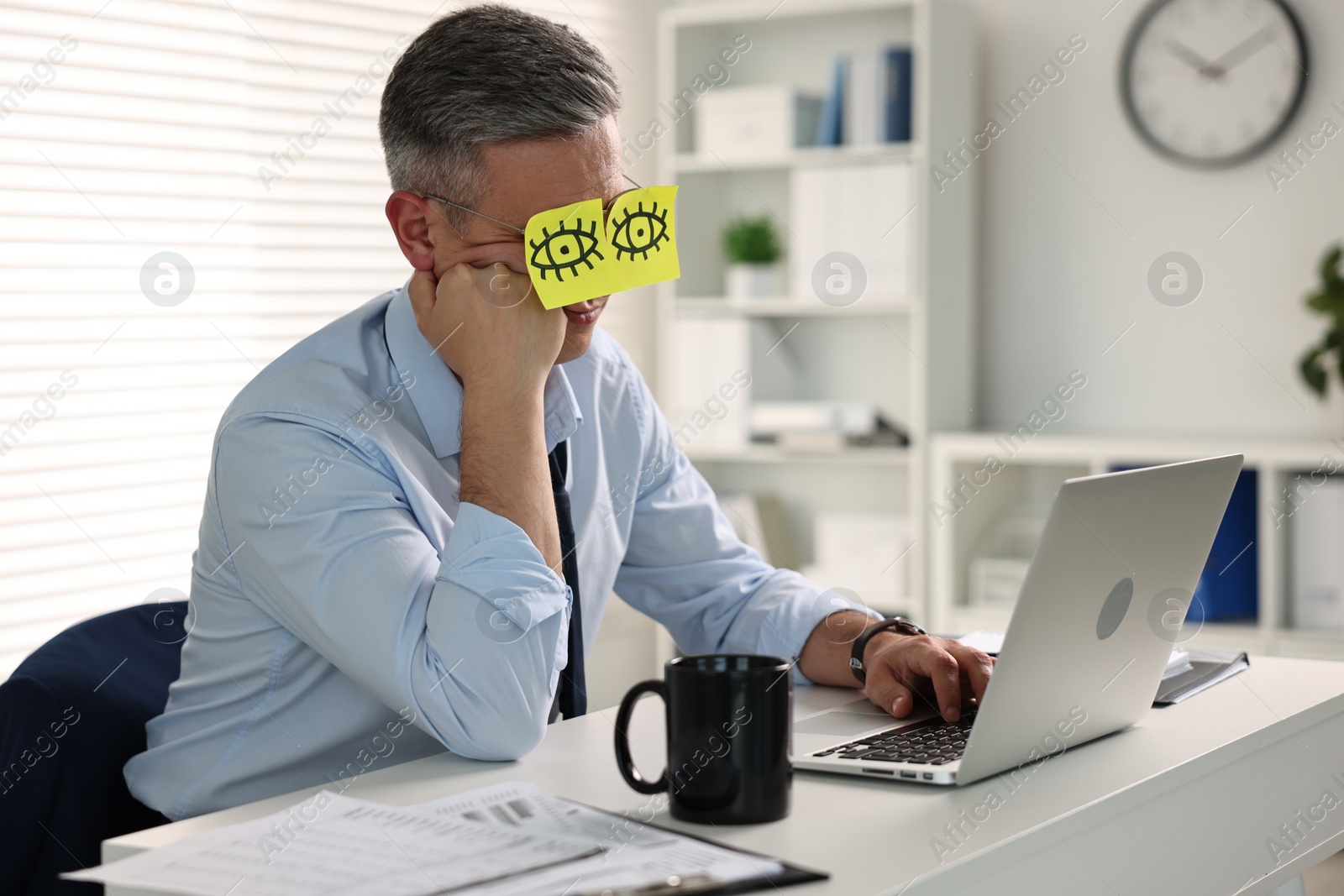 Photo of Man with fake eyes painted on sticky notes snoozing at workplace in office