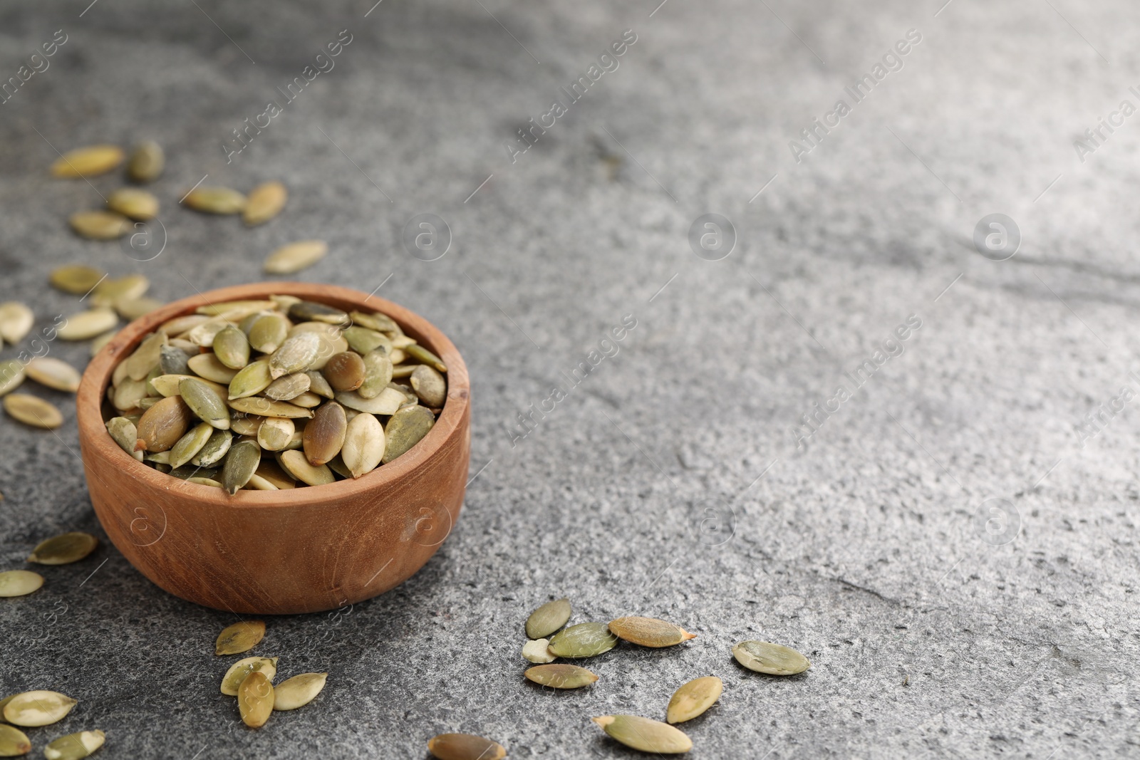 Photo of Wooden bowl with peeled pumpkin seeds on grey table. Space for text