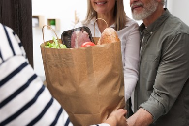 Photo of Courier giving paper bag with food products to senior couple indoors, closeup