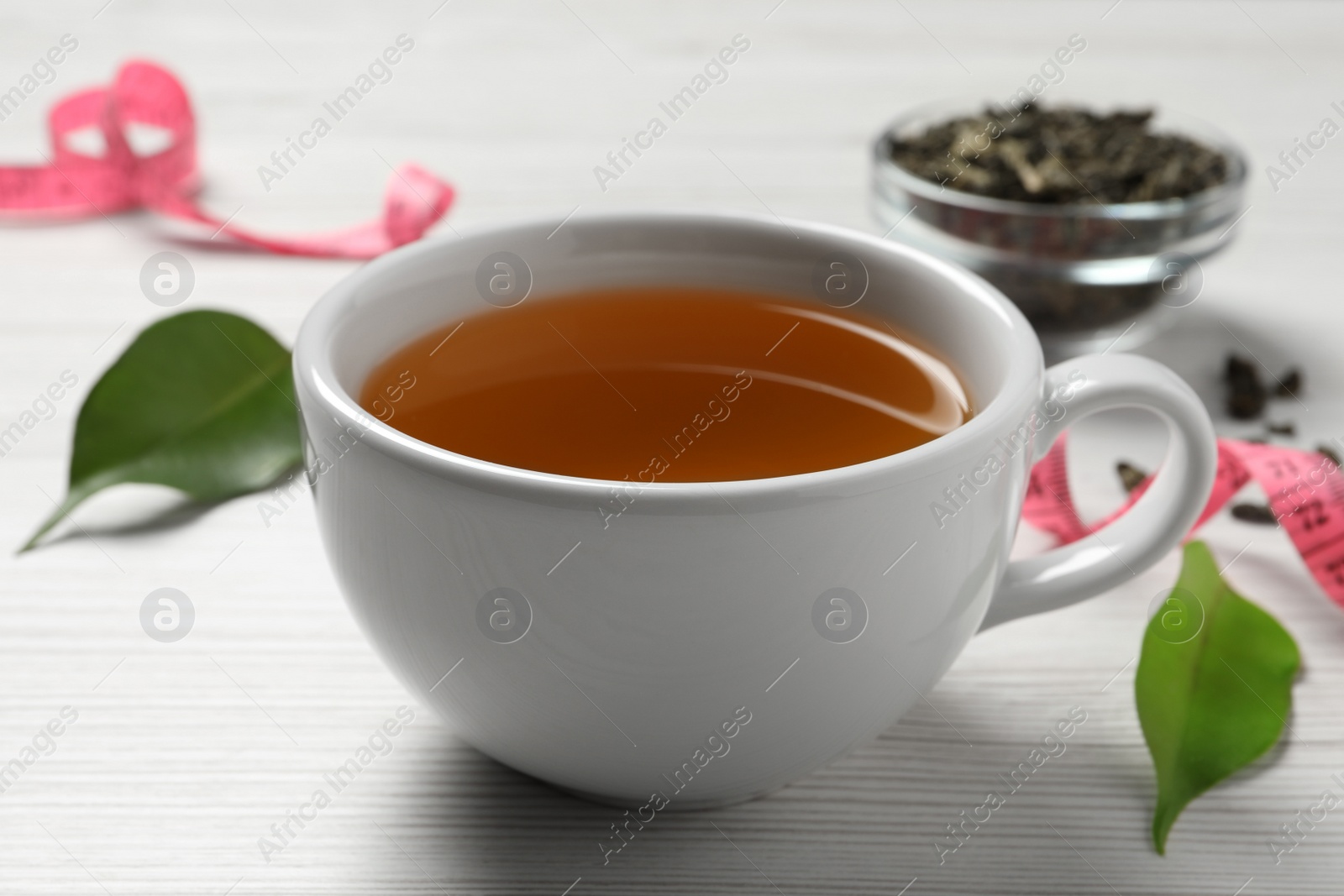 Photo of Cup of herbal diet tea, measuring tape and green leaves on white wooden table, closeup. Weight loss concept