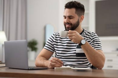 Photo of Young man with cup of coffee watching webinar at table in room