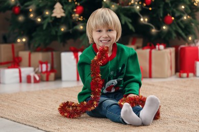 Photo of Little child with bright tinsel on floor at home. Christmas decoration