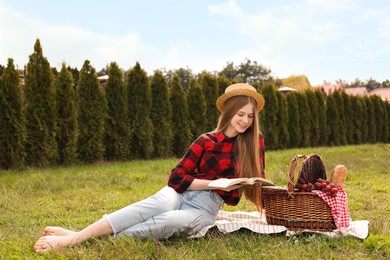 Happy girl with picnic basket reading book on green grass in park