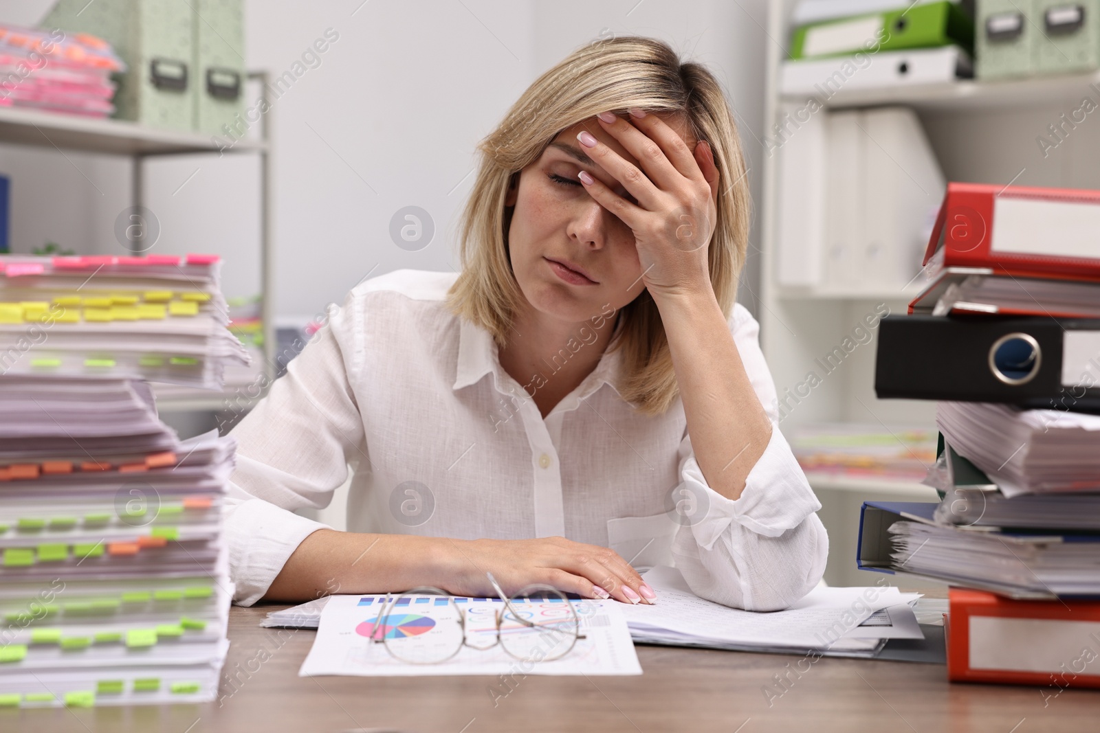 Photo of Overwhelmed woman sitting at table with stacks of documents and folders in office